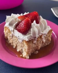 A slice of pink strawberry tres leches cake topped with whipped cream and fresh strawberries on a plate