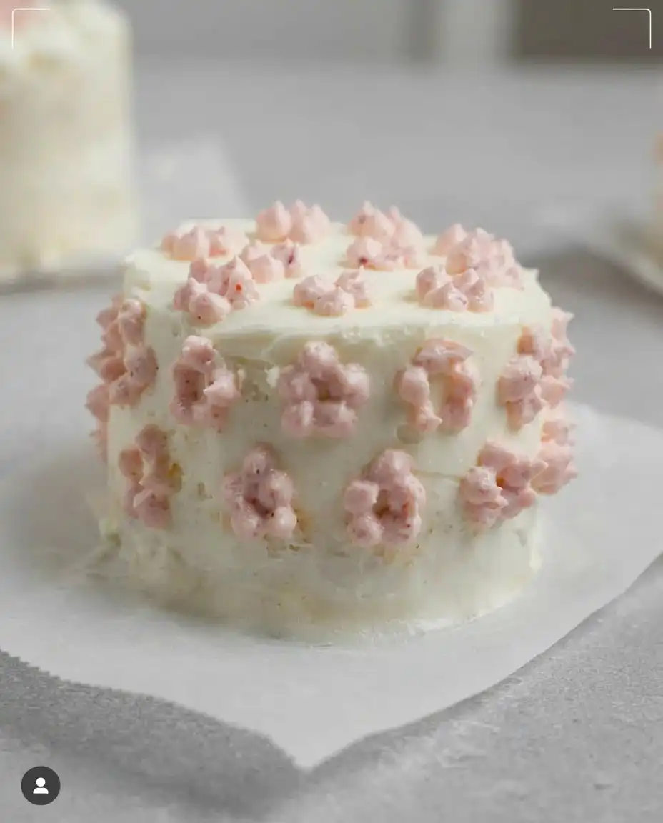 A small vanilla cake with purple decorations nestled inside a white clamshell container