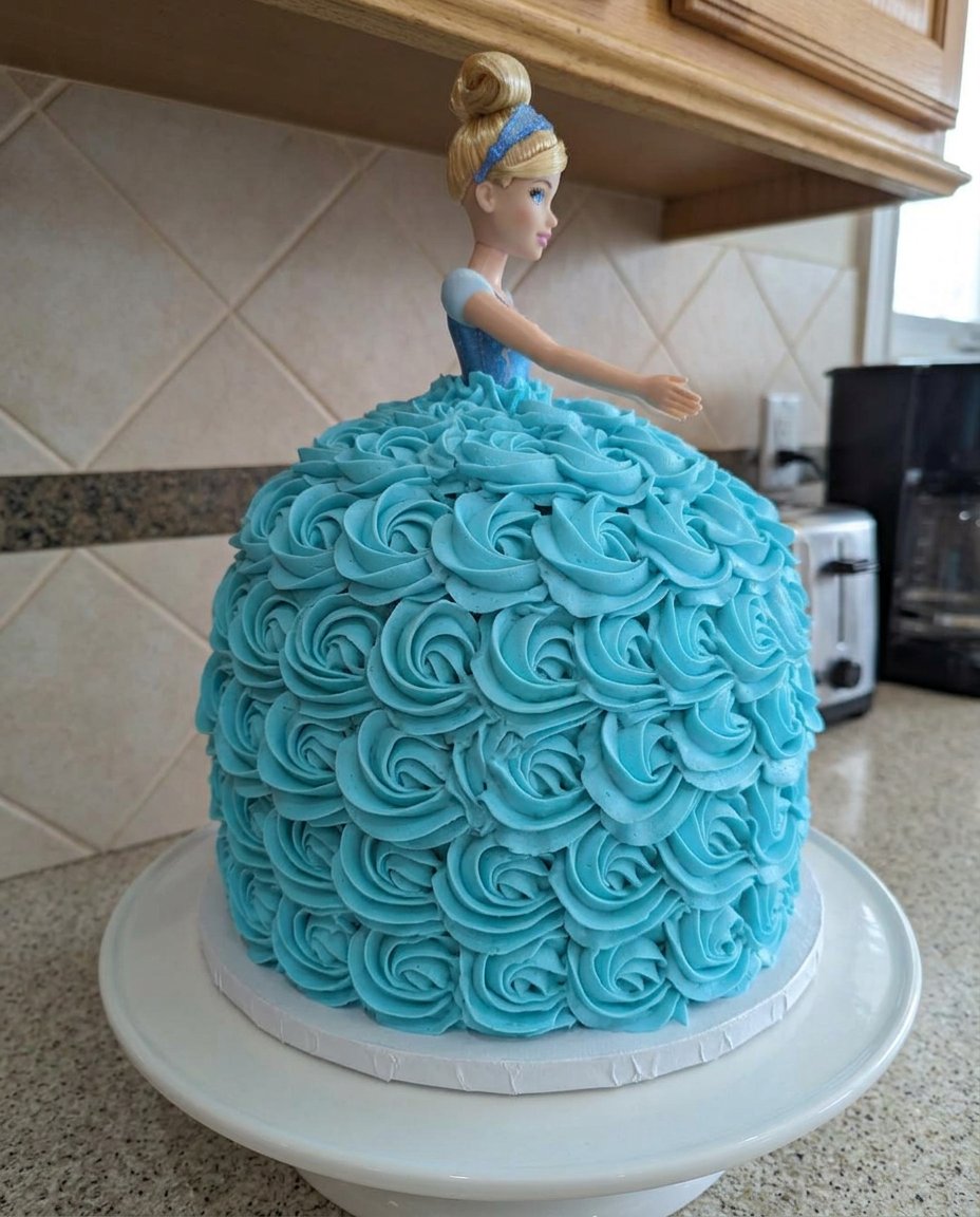 A completed Cinderella doll cake with blue frosting rosettes on a vintage stand