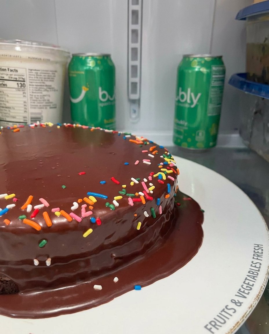Warm chocolate ganache being poured over a dark chocolate cake layer on a wire cooling rack.