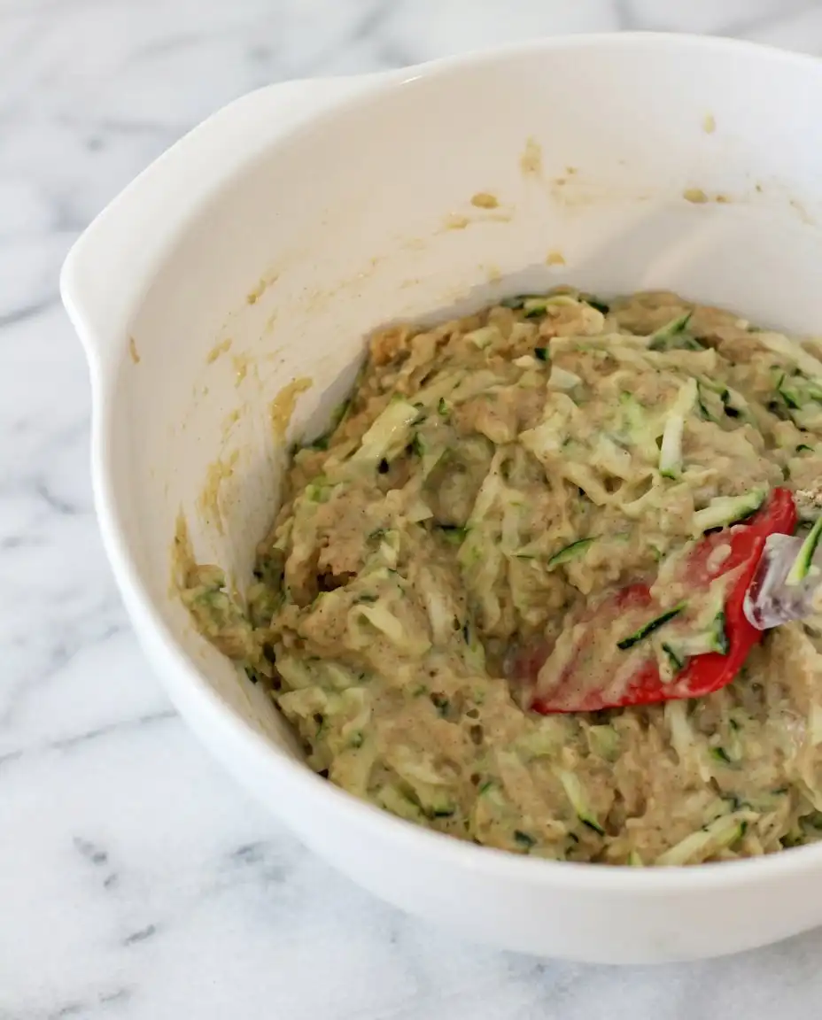 A wooden spoon folding shredded zucchini into Chocolate Chip Zucchini Muffins batter.