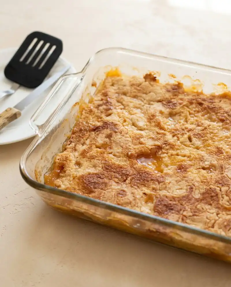 A golden Peach Cobbler with Yellow Cake Mix resting in a glass baking dish on a wooden table.