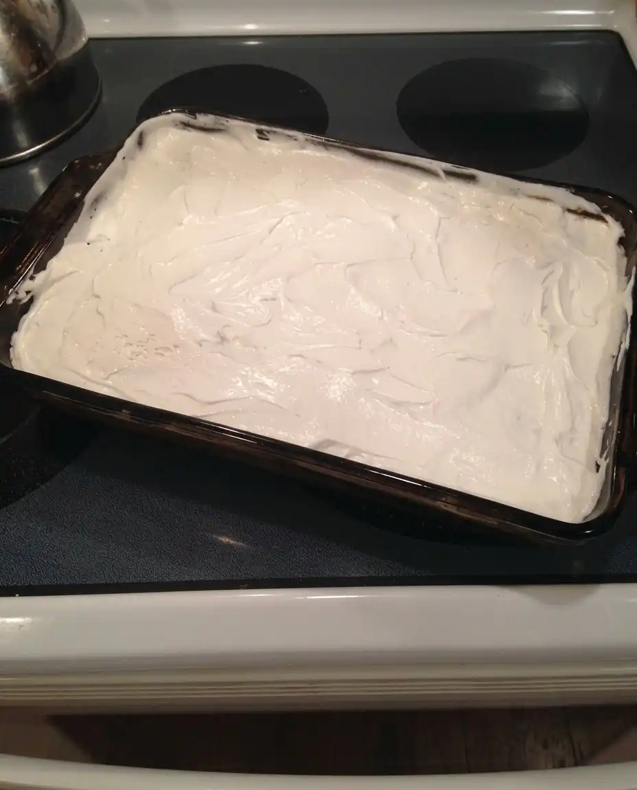 A hand using an offset spatula to spread a smooth layer of chocolate pudding over the cream cheese layer
