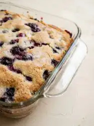 A golden brown slice of Blueberry Buttermilk Breakfast Cake showing juicy berries and a crackly sugar crust.