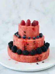 A stunning three-tiered Watermelon and Berry Layer Cake decorated with peaches and blueberries on a marble platter